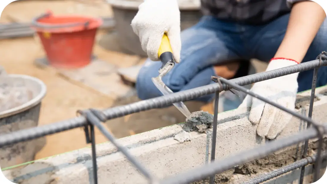 Gloved hands trowelling mortar along a concrete block wall with reinforcing bar set behind it.