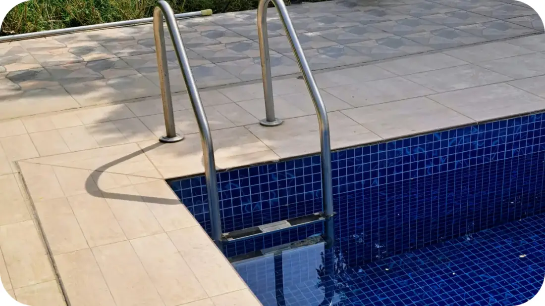 Corner of a pool finished in deep-blue mosaic tile with stainless handrails and paved decking.