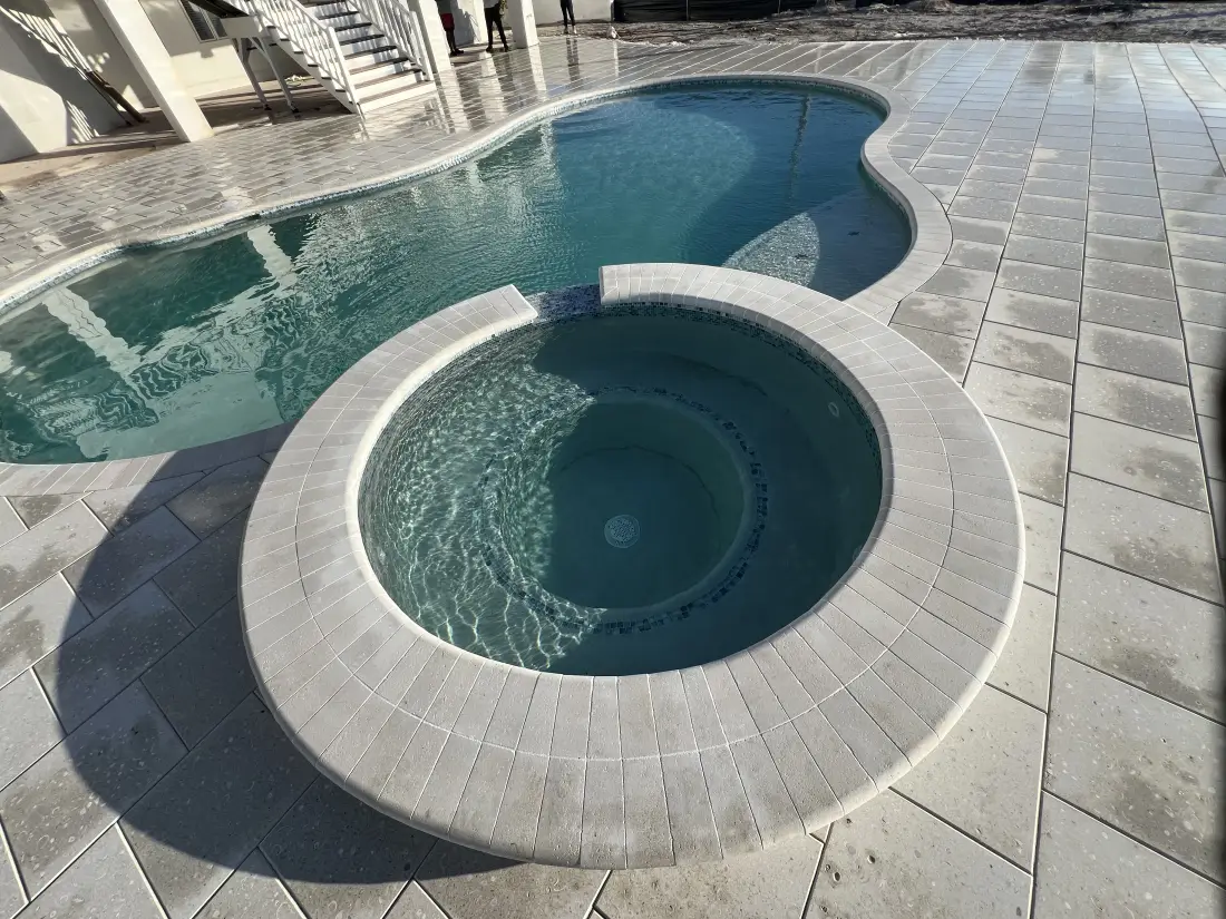 Close view of a round raised spa built into travertine paver decking, ocean visible behind.