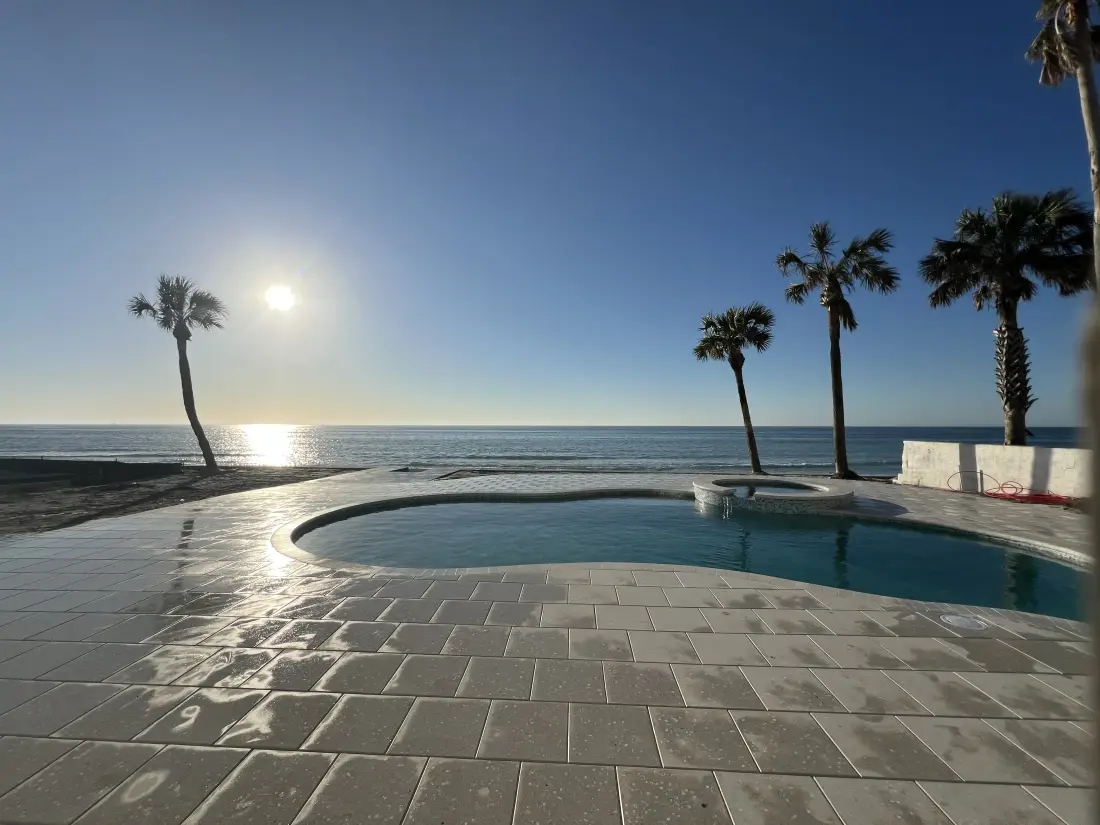 Freeform beachfront pool at sunset with silhouetted palm trees.