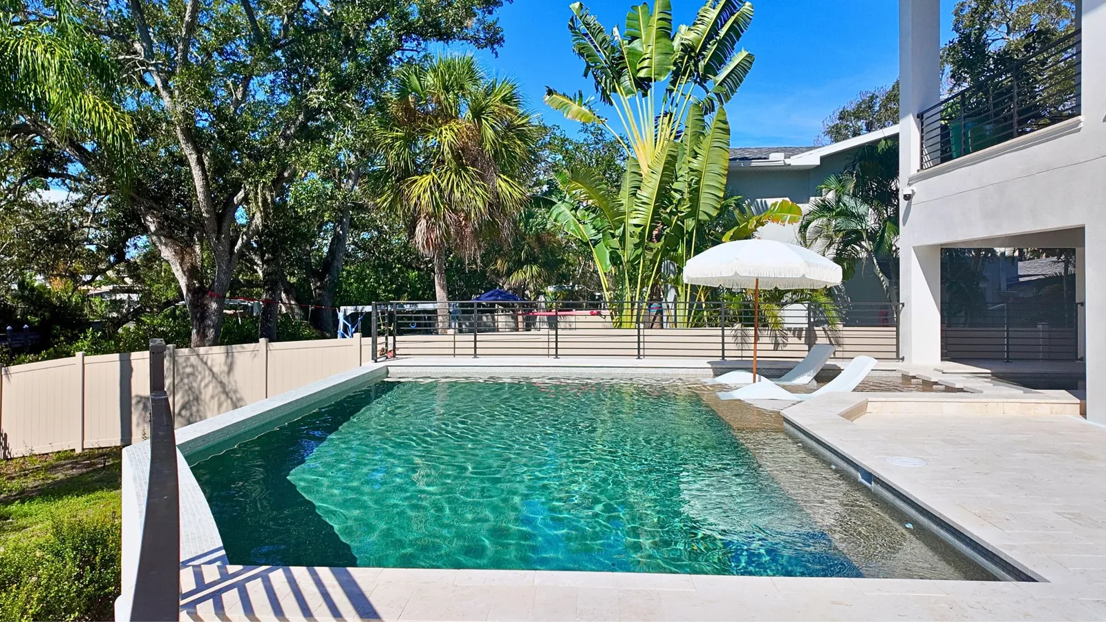The full length of the Belmar Gardens pool seen from the lawn, its sunshelf and fringed umbrella set against the white two-story house and a stand of live oaks and palms.