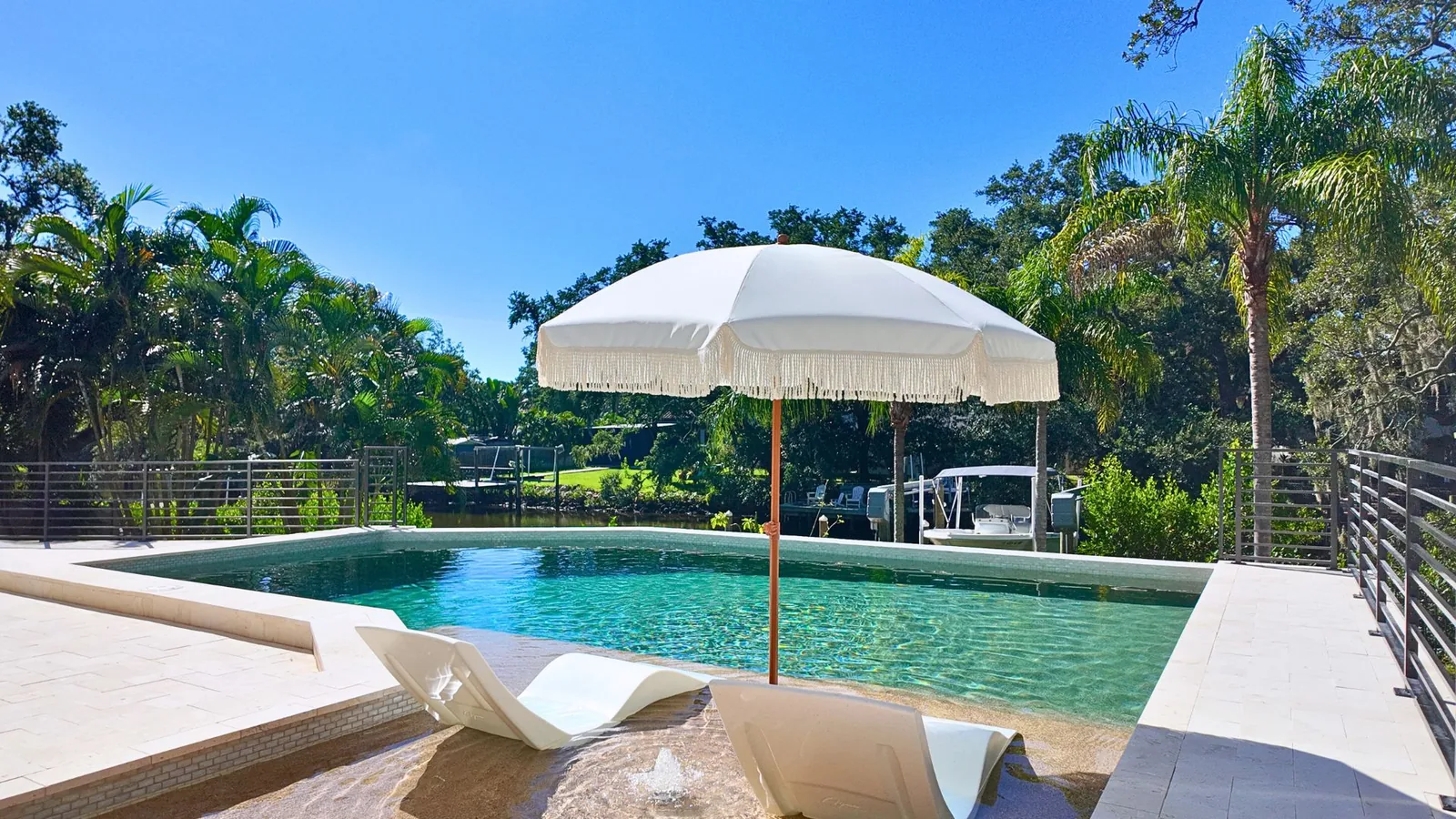 In-water loungers and a fringed umbrella on the sunshelf, with the cable railing and a boat dock on the water beyond.