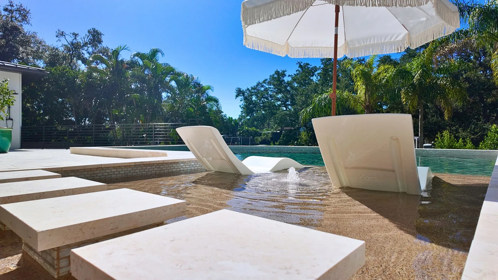 Square stepping stones crossing the shallow sunshelf, two in-water loungers behind them and the bubbler running between.