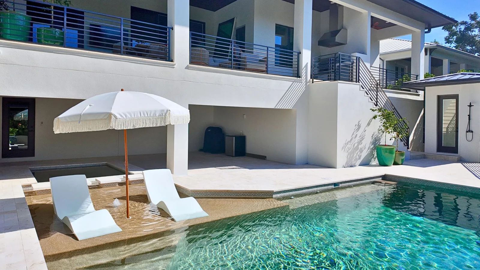 Two loungers standing in the shallow sunshelf under a fringed umbrella, the tiled spa directly behind them and the house's balcony above.