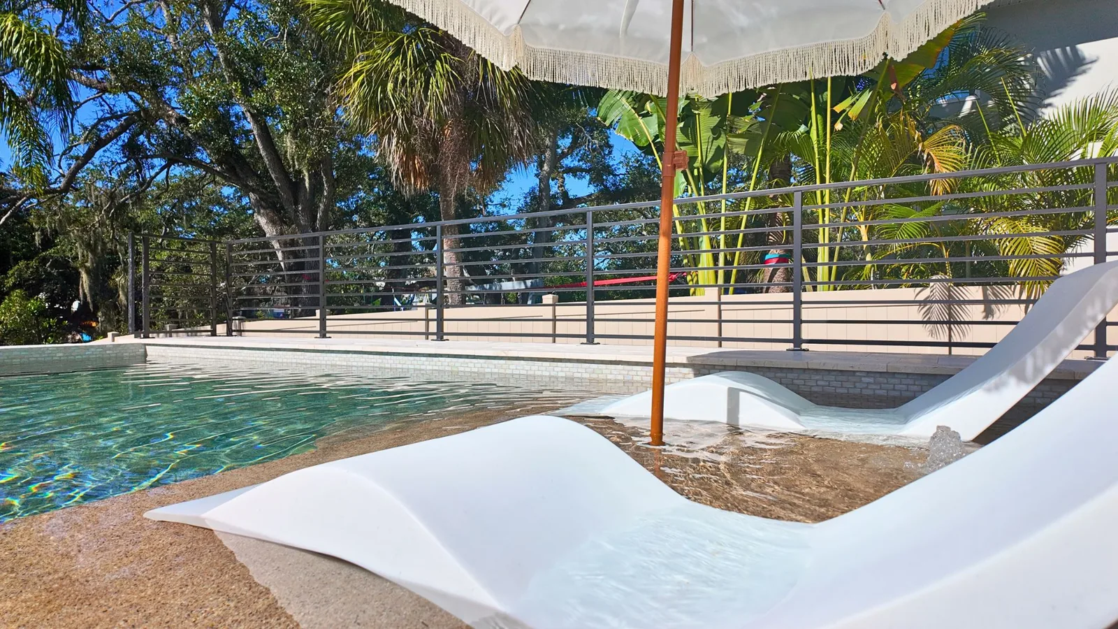 A lounger sitting in the shallow water of the sunshelf, the umbrella pole and cable railing framing the oaks beyond.