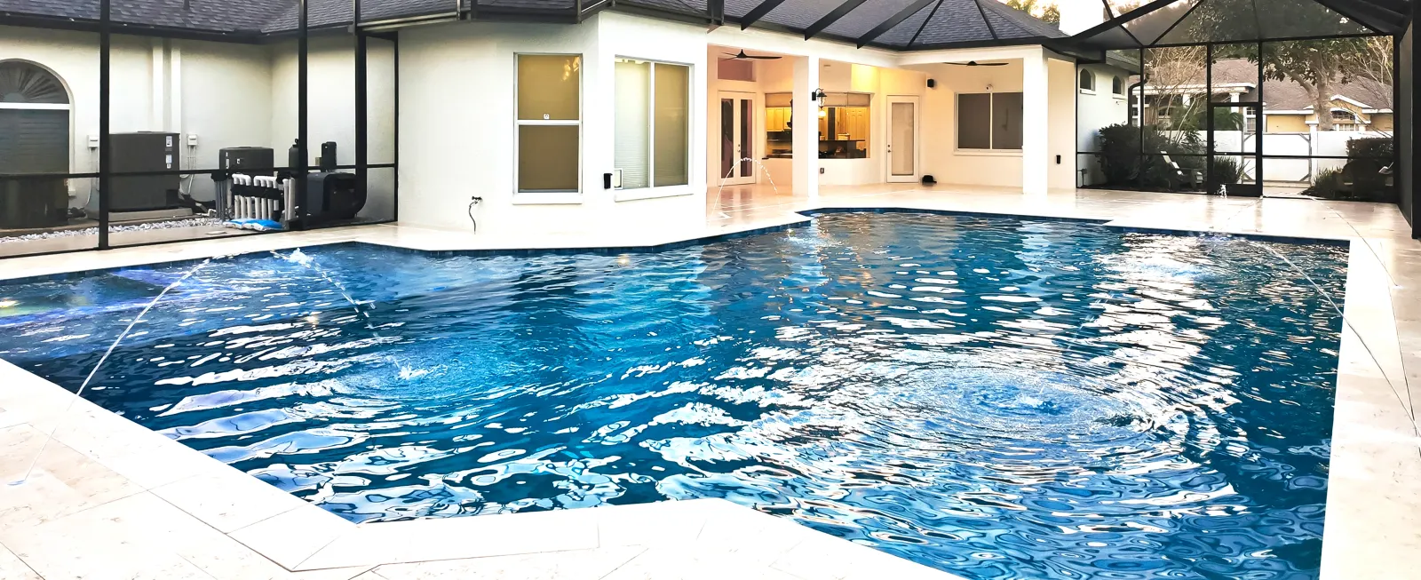 Rectangular pool with dark blue tile inside a screened enclosure, home lit up behind at dusk.