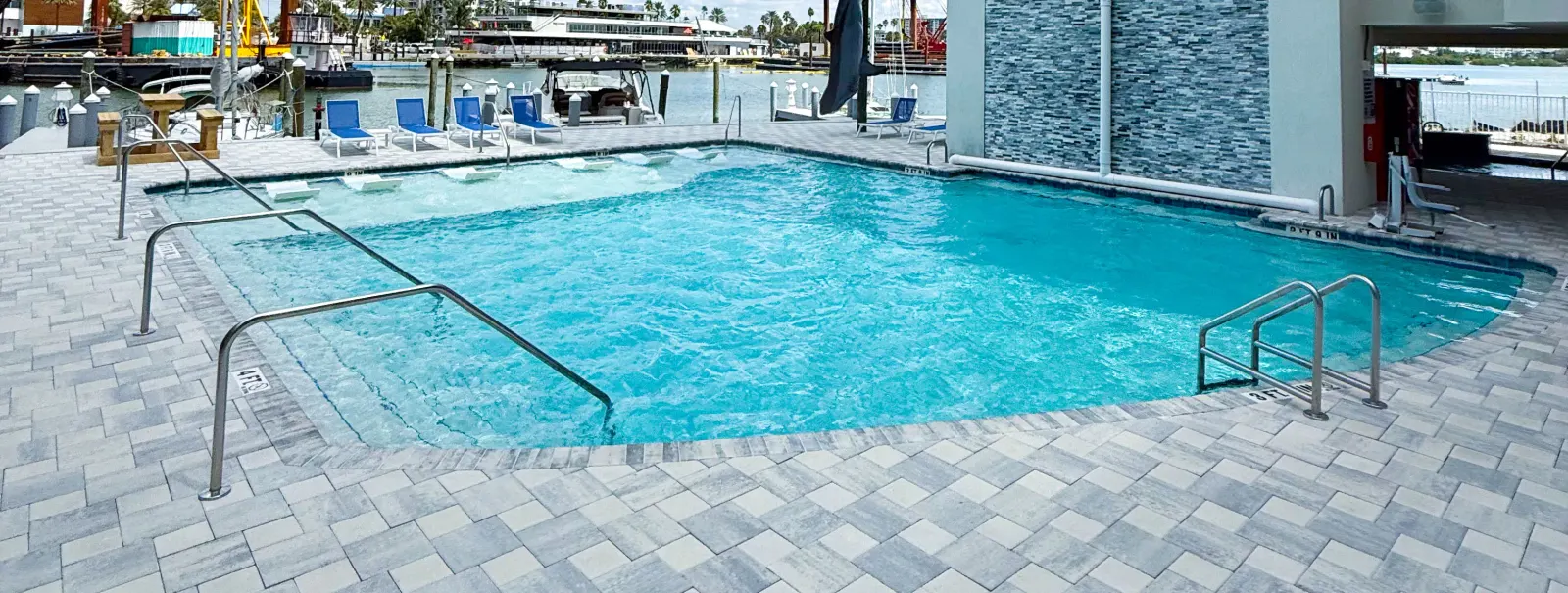 Rectangular pool deck beside a marina, lounge chairs facing the water, a multi-story building behind.