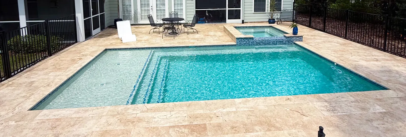 Rectangular pool with an attached raised spa and travertine decking, patio furniture on a residential lot.
