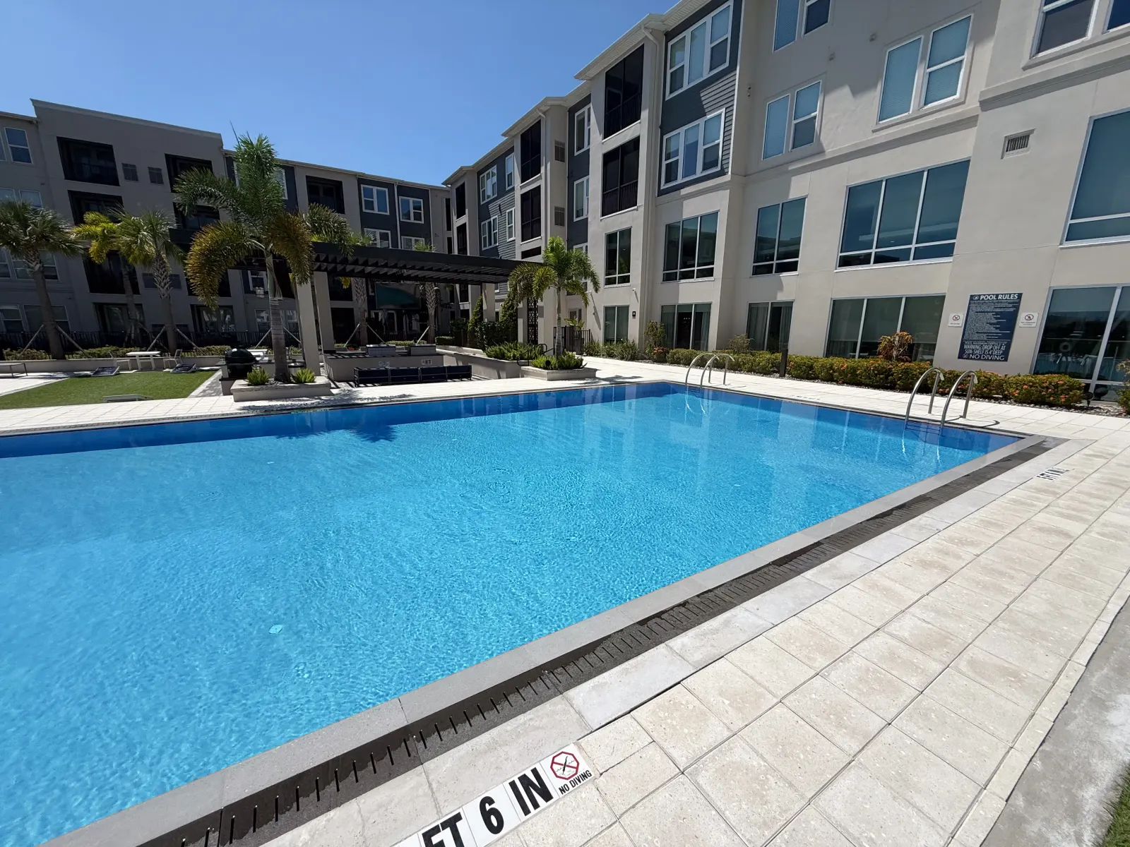 Large rectangular community pool alongside a multi-story residential building, palm trees along the deck.