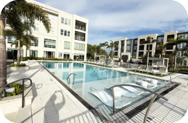 Pool deck with handrails and lounge seating beside the apartment building, palm trees around the courtyard.
