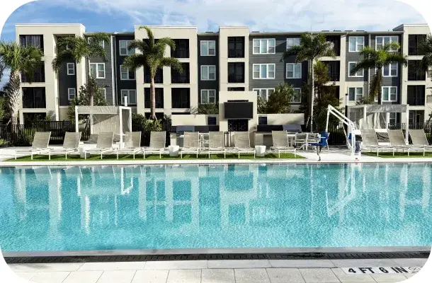 Wide view of the community pool and deck furniture with the residential building behind.