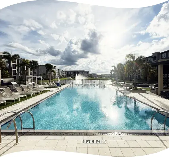 Wide view of the pool deck with a fountain jet and surrounding apartment buildings.
