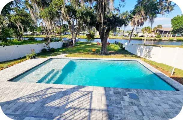 Finished rectangular pool with paver decking, oak trees, and a pond visible beyond the fence.