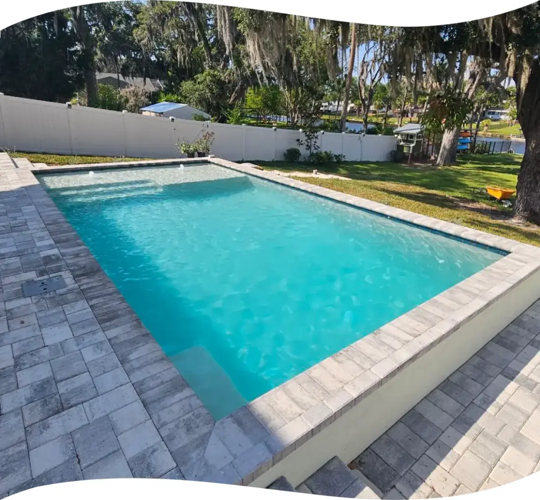 Finished rectangular pool with paver decking in a fenced, tree-lined yard.