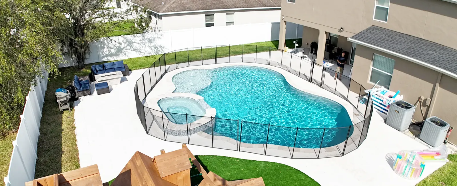 Aerial view of a freeform lagoon-shaped pool with an attached spa and a child's playset in the fenced backyard.