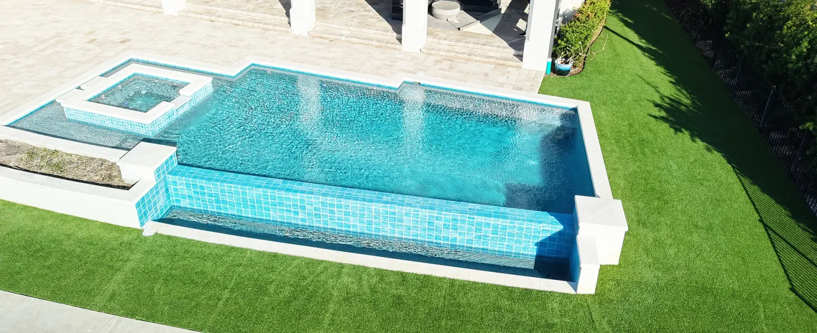 Aerial view of a long rectangular pool with a raised spa and water jets on a manicured lawn.