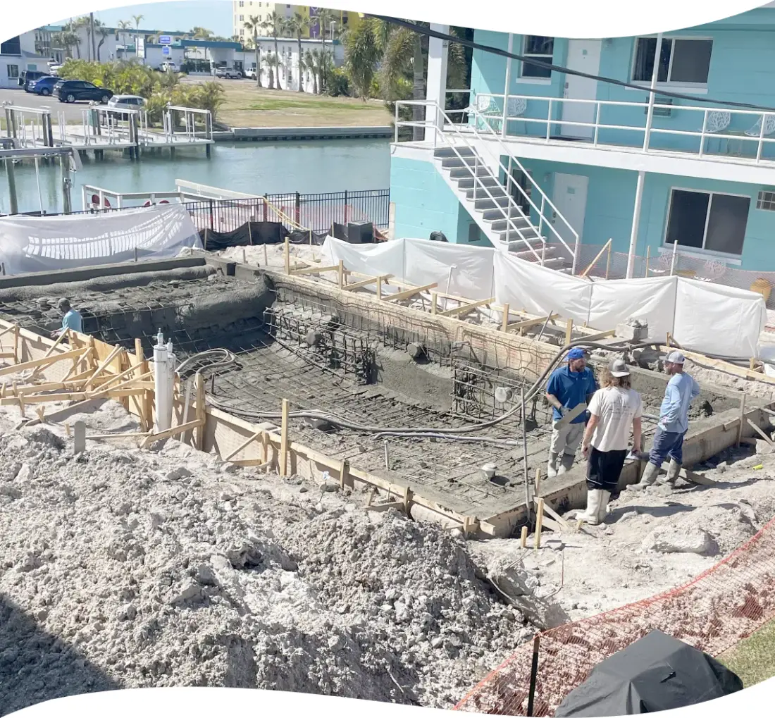 Pool under construction beside a canal: an open excavation with timber forming, a rebar cage and grade stakes, crew on site.