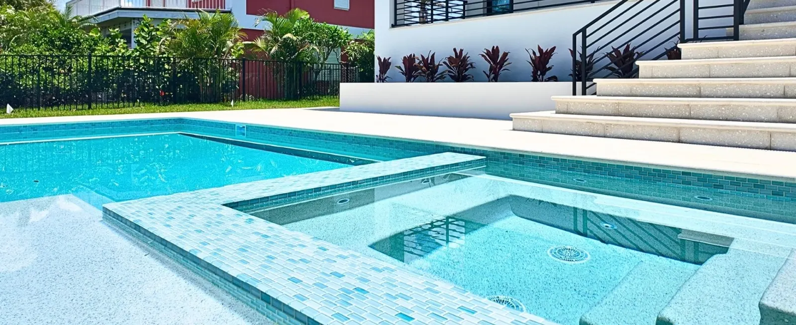 Modern two-story home with a rectangular pool, raised spa, and sun-shelf steps, framed by a black metal safety fence and tropical landscaping.