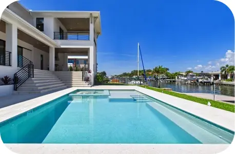 Wide view of a two-story waterfront home with pool and spa alongside a canal lined with docked boats.