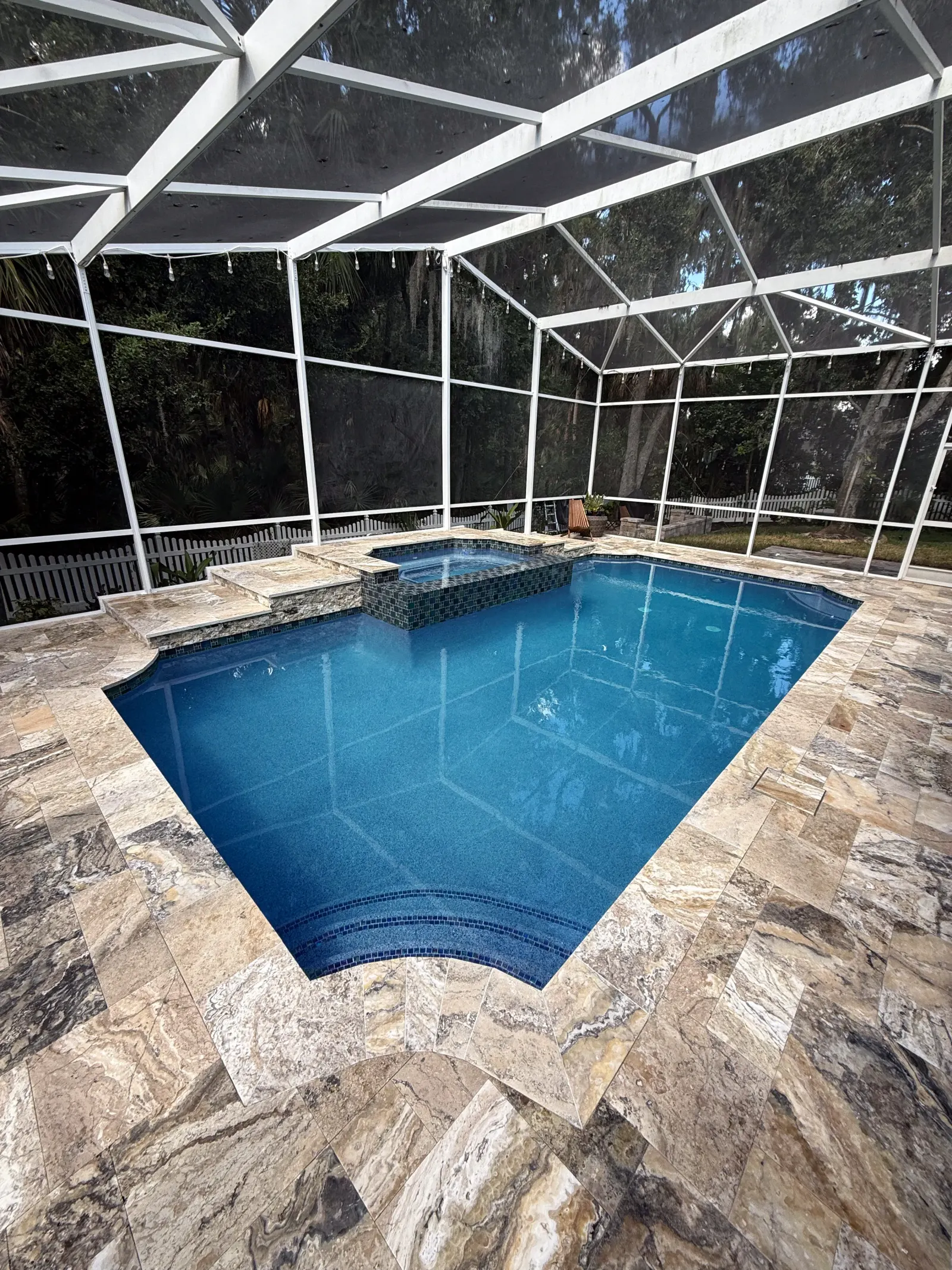 Elevated view of a geometric pool with angled corners and travertine decking on a wooded lot.