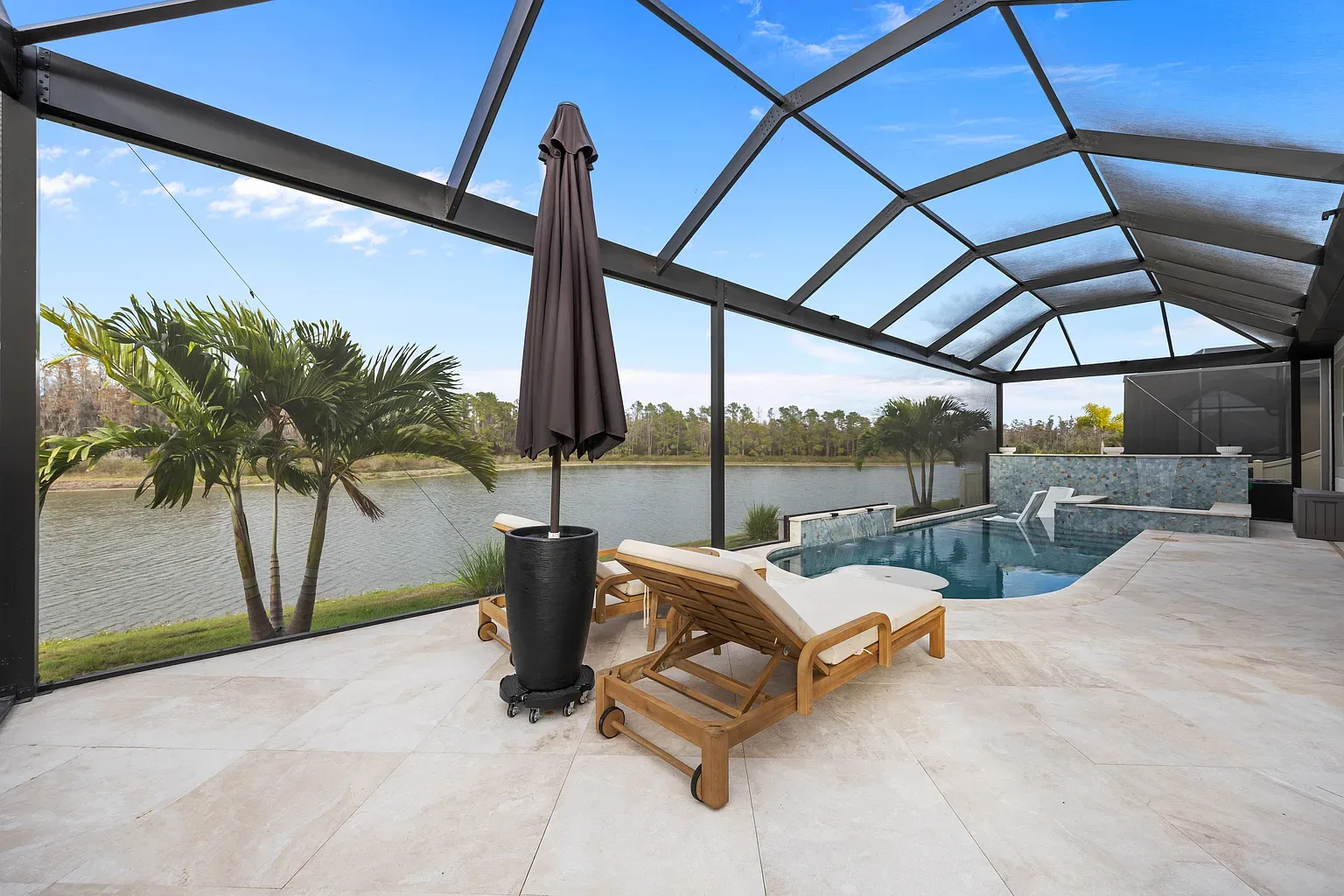 Teak loungers and a folded umbrella on the travertine pool deck of a screened lanai overlooking a lake in Wesley Chapel, Florida.