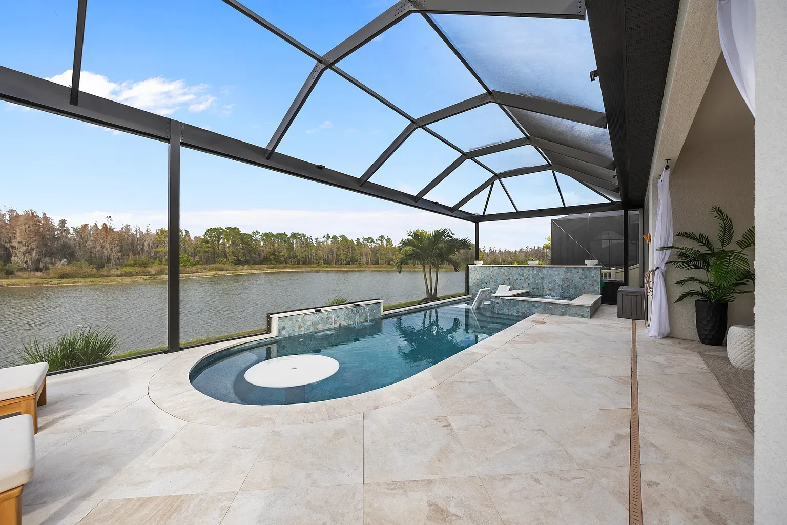 Narrow linear pool and raised spa with a white in-pool table and sheer descent waterfalls, inside a screened lanai on a lake in Wesley Chapel, Florida.
