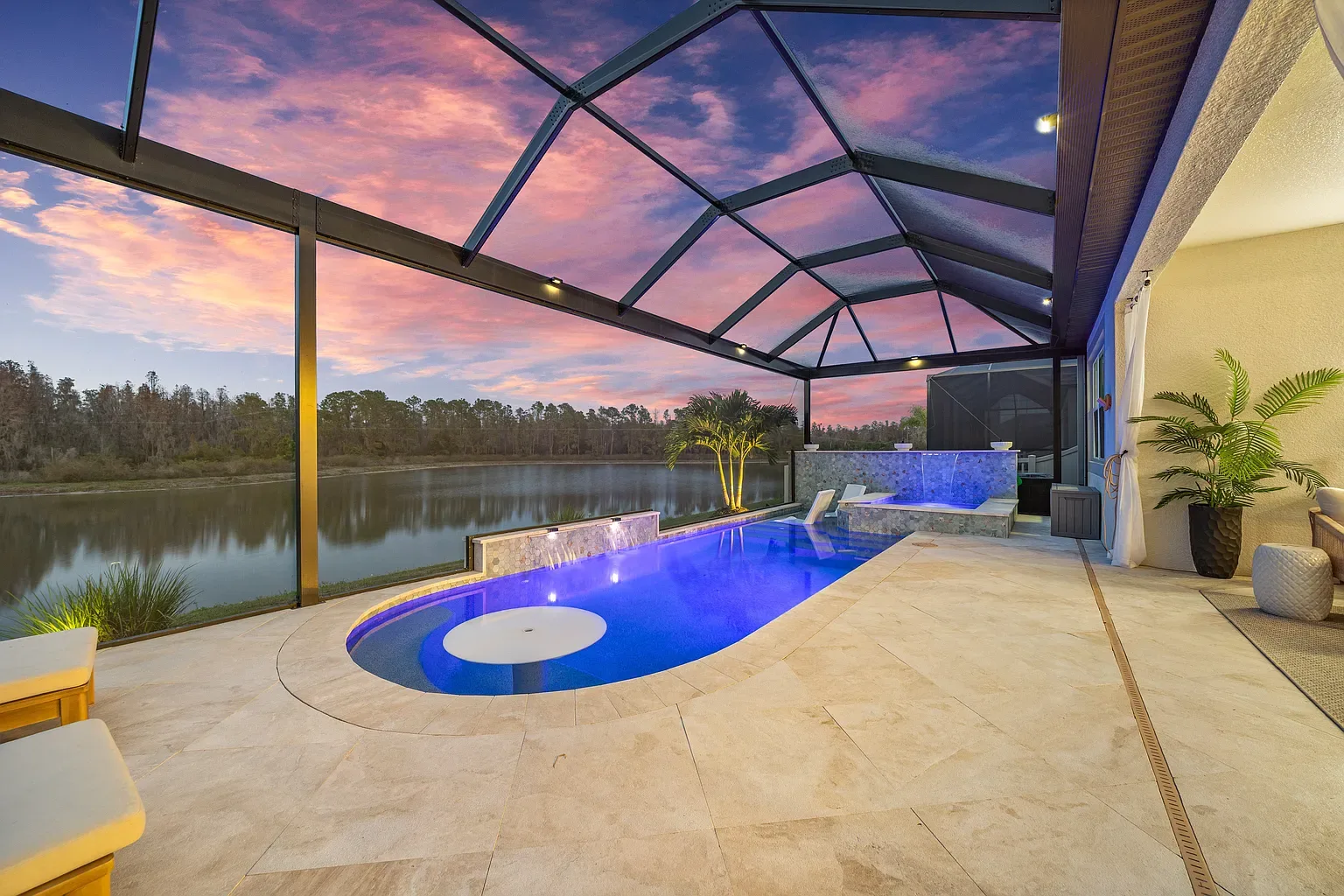 Linear pool lit blue by its LED lighting at dusk, raised spa and lake behind the screen enclosure, Wesley Chapel, Florida.